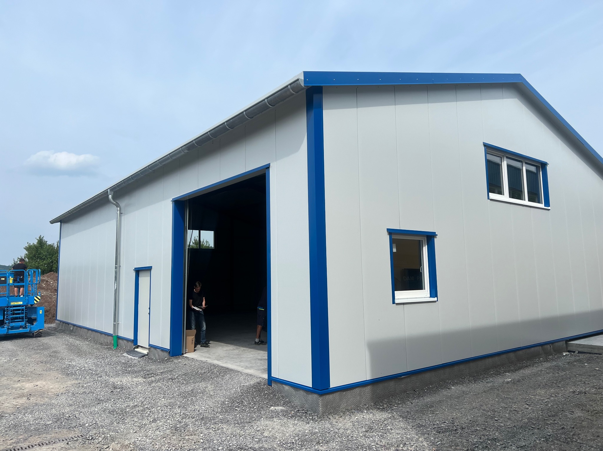 Gotowy budynek z płyt warstwowych, szare ściany z niebieskimi obramowaniami, widoczne okna i otwarte wrota, żwirowe podłoże, na zewnątrz osoby i podnośnik.