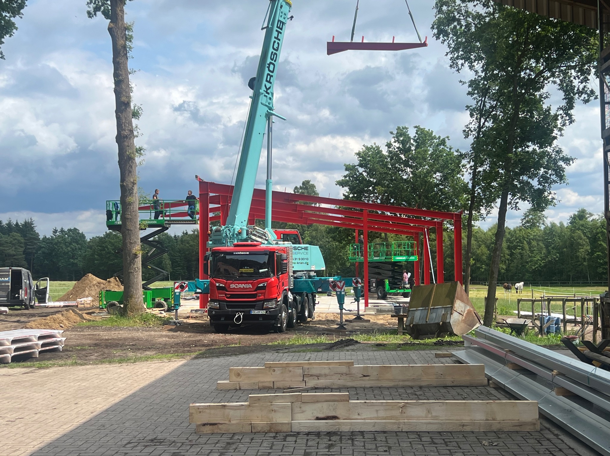 Montaż konstrukcji stalowej: czerwony szkielet budynku, podnośniki, dźwig Krösche, elementy konstrukcyjne na ziemi. Prace w toku, widok z bliska na plac budowy.