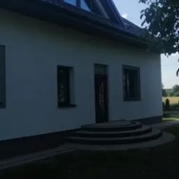 Boczny widok domu jednorodzinnego z jasną elewacją, ciemną stolarką okienną i drzwiami wejściowymi, trójkątne okno na poddaszu, schody wejściowe wyłożone kostką, drzewo w prawym górnym rogu.