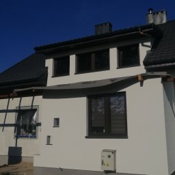 FHU Błazej Szawłowski - Fragment elewacji domu jednorodzinnego w trakcie prac wykończeniowych, z widocznym zabezpieczeniem okien folią i elementami rusztowania, świeżo pomalowana ściana oraz osłona przeciwsłoneczna...