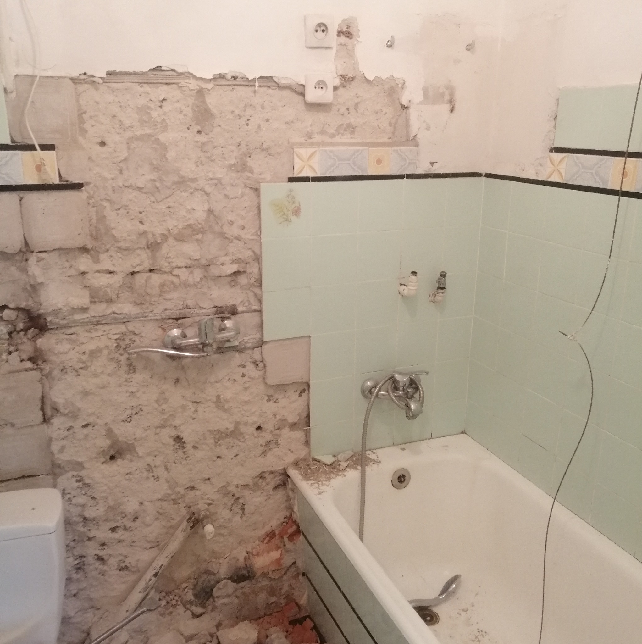 Łazienka w trakcie remontu: odsłonięta cegła, fragmenty starych płytek, biała wanna, zdemontowane elementy instalacji wodnej, widoczne gniazdka elektryczne na ścianie.