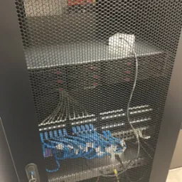 Serwerownia z widocznymi niebieskimi kablami sieciowymi, czarną obudową i kluczykiem do szafy rack na drzwiach.