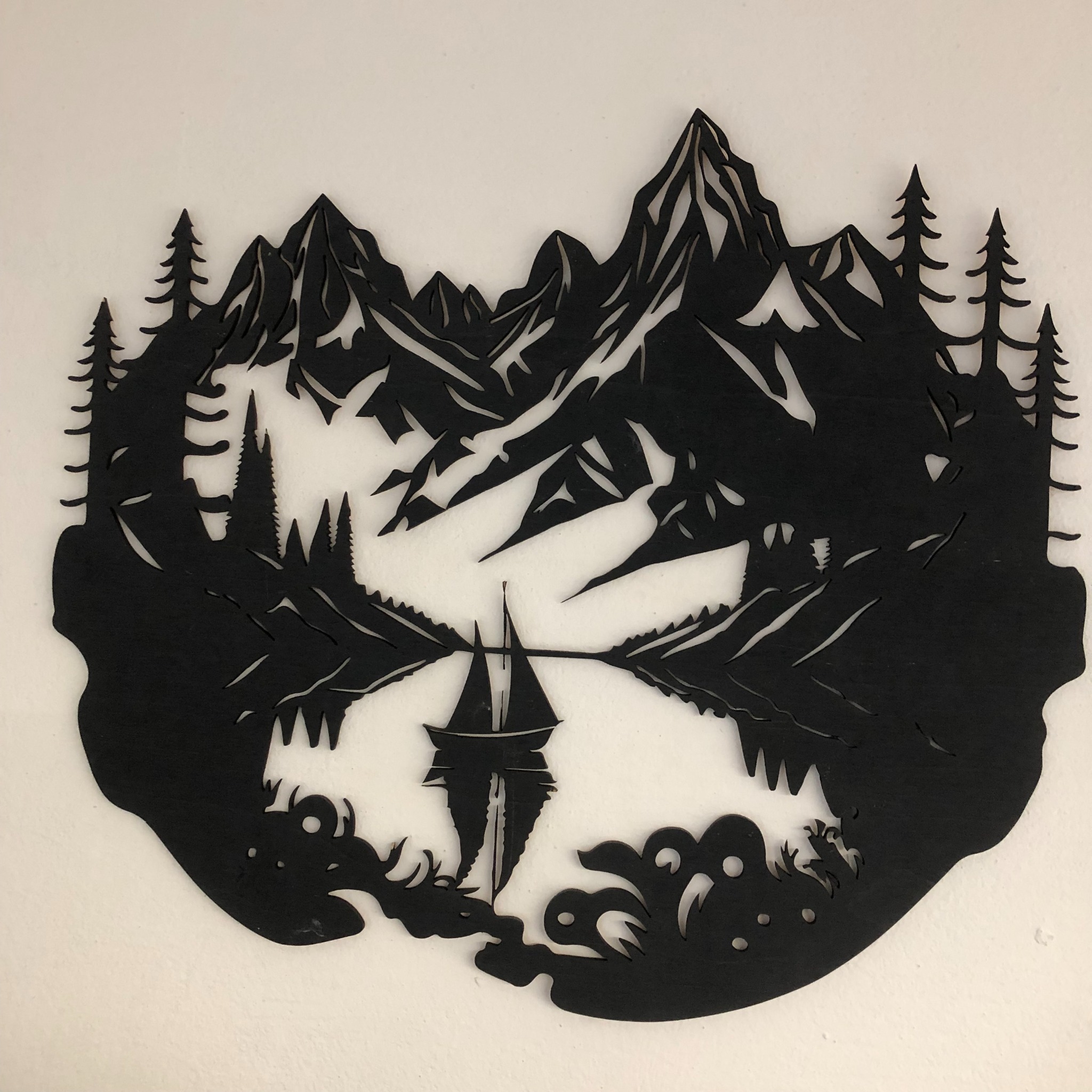 Wycięty laserowo, czarny, drewniany obraz przedstawiający górski krajobraz z jeziorem i żaglówką, wiszący na białej ścianie.