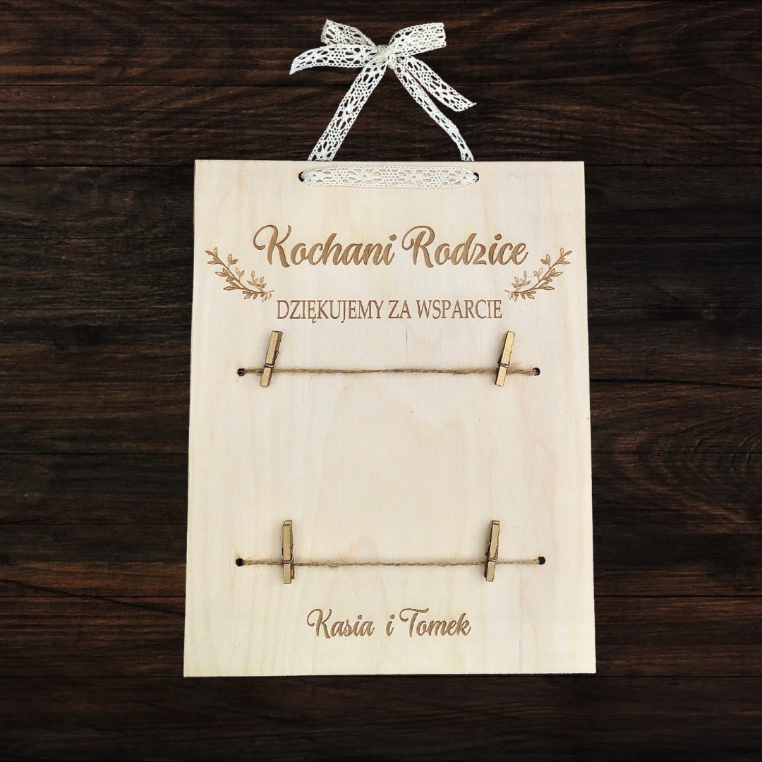 Drewniana tablica z grawerem 'Kochani Rodzice Dziękujemy za wsparcie' i imionami 'Kasia i Tomek', zawieszona na sznurkach z klamerkami, ozdobiona koronkową kokardką.