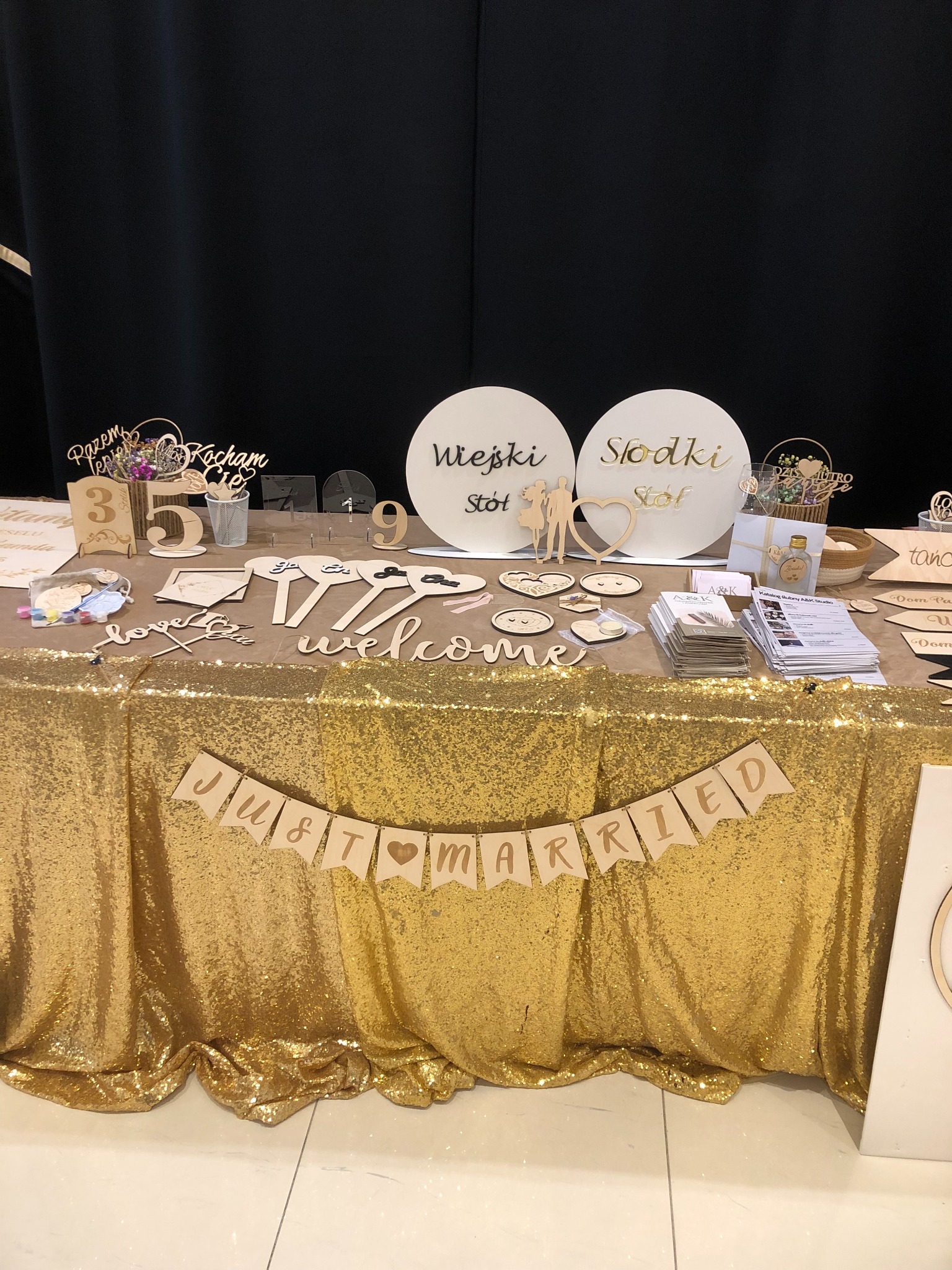 Dekoracje ślubne z drewna na złotym, błyszczącym obrusie: napisy 'Wiejski Stół', 'Słodki Stół', 'Just Married', numery stołów, serca z uśmiechami, ulotki reklamowe.