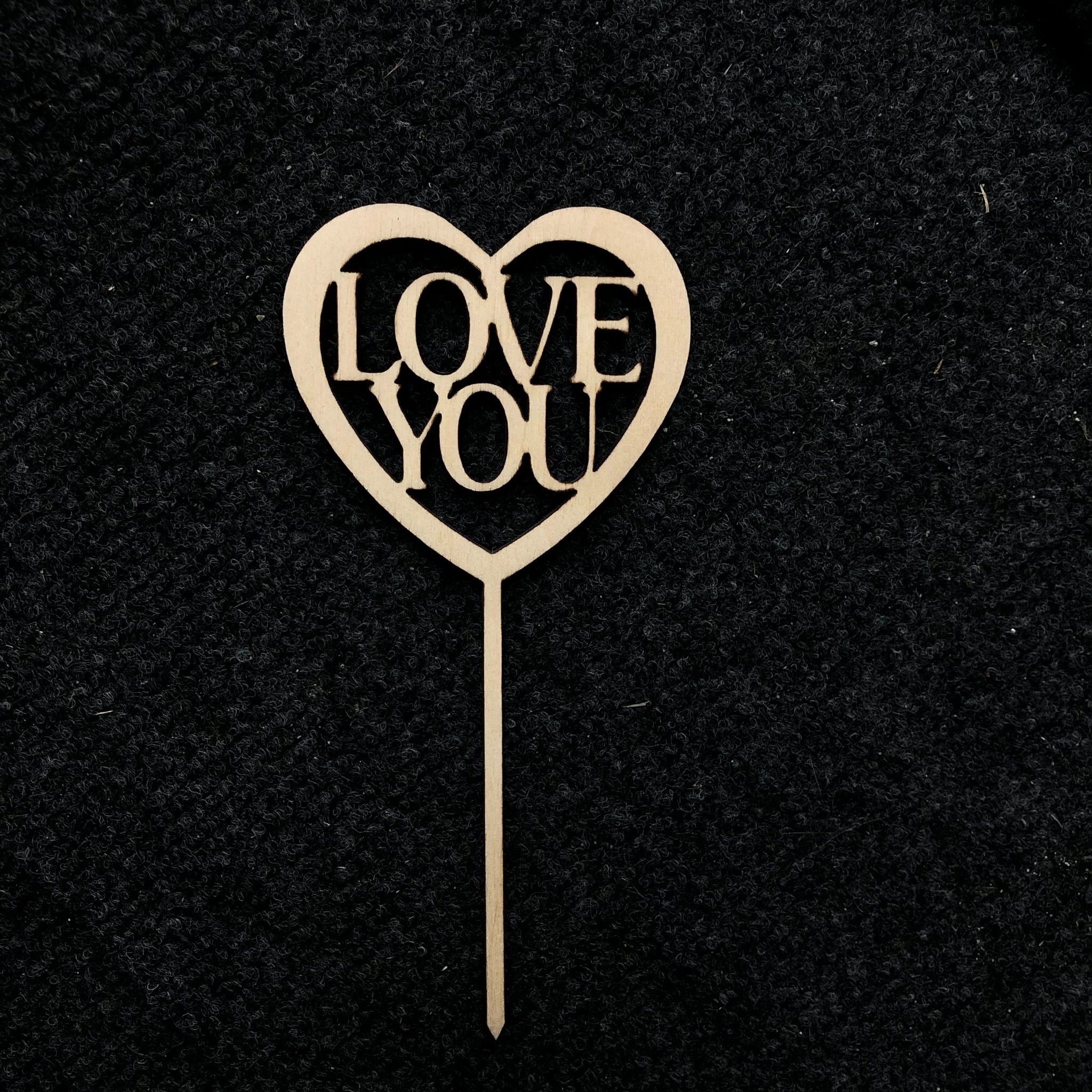 Drewniany topper na tort w kształcie serca z napisem 'LOVE YOU', wykonany techniką CNC, leżący na czarnym, teksturowanym tle.