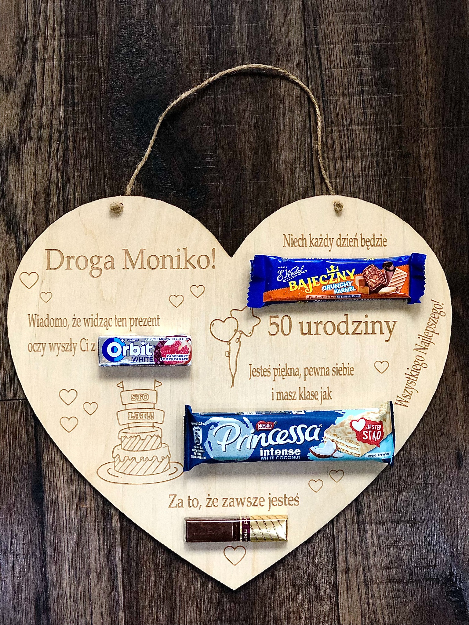 Drewniane serce z wygrawerowanymi życzeniami urodzinowymi i przymocowanymi słodyczami, leżące na drewnianym tle.
