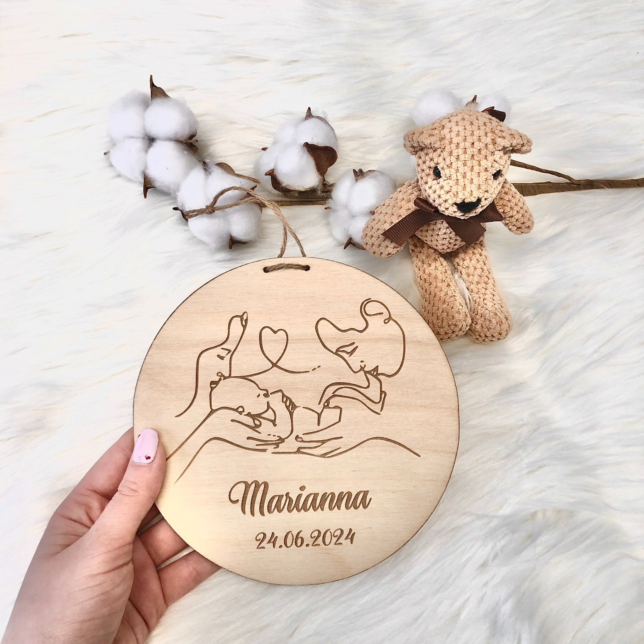 Drewniana zawieszka z wygrawerowanym imieniem Marianna i datą 24.06.2024, przedstawiająca stylizowany rysunek matki trzymającej dziecko, z dekoracją z bawełny i pluszowym misiem na puszystym białym...