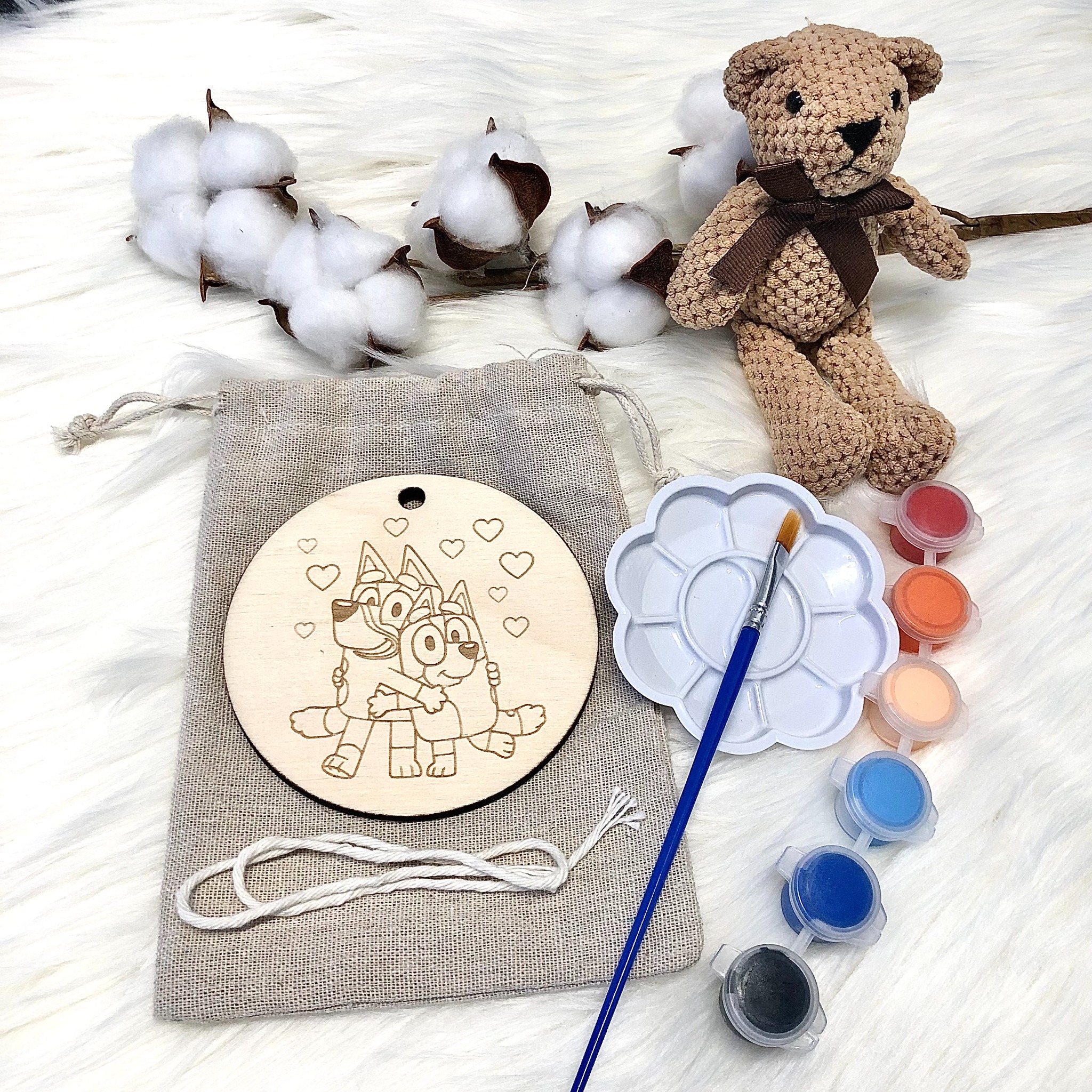 Drewniana zawieszka z wygrawerowanymi postaciami z bajki Bluey, leżąca na lnianym woreczku obok farb, pędzla, pluszowego misia i gałązek bawełny na białym futrzanym tle.