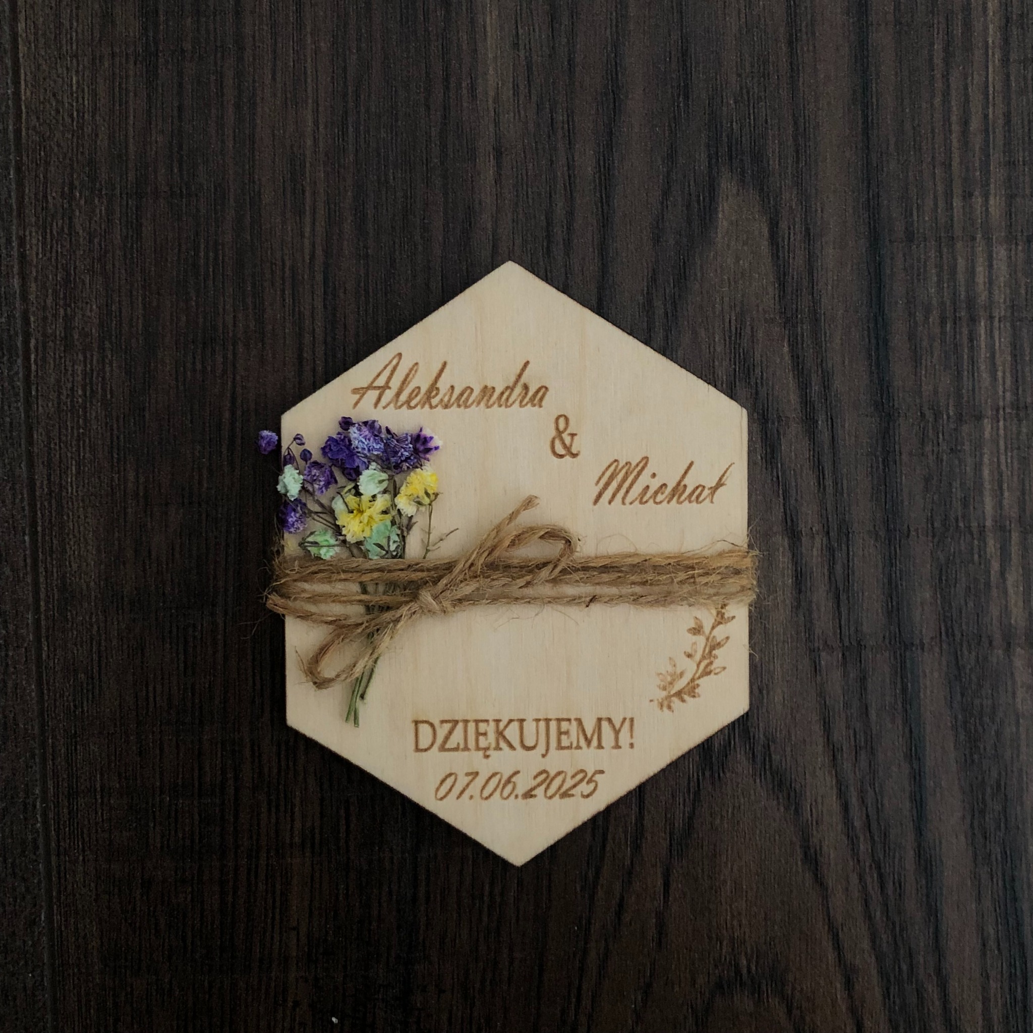 Drewniana zawieszka w kształcie sześciokąta z wygrawerowanymi imionami Aleksandra i Michał, datą 07.06.2025 i napisem Dziękujemy, ozdobiona suszonymi kwiatami i jutowym sznurkiem na tle ciemnego...