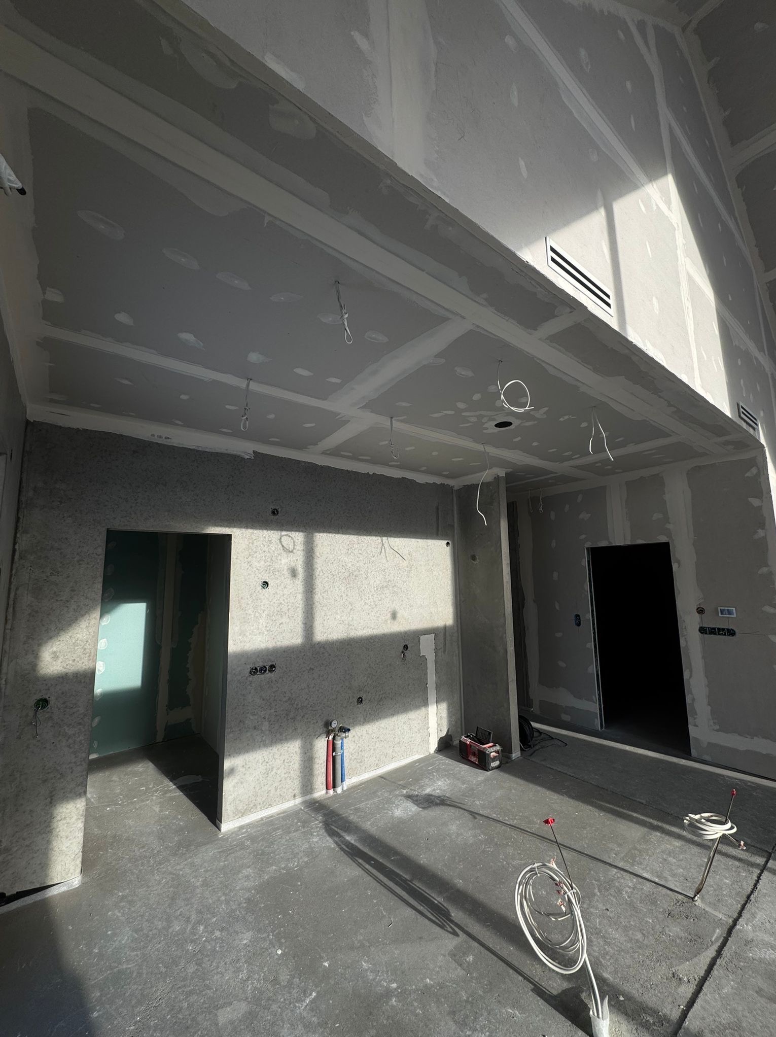 Surowe wnętrze w trakcie wykańczania: płyty karton-gips na ścianach i suficie, widoczne instalacje, otwory drzwiowe, wentylacja. Wnętrze doświetlone naturalnym światłem.