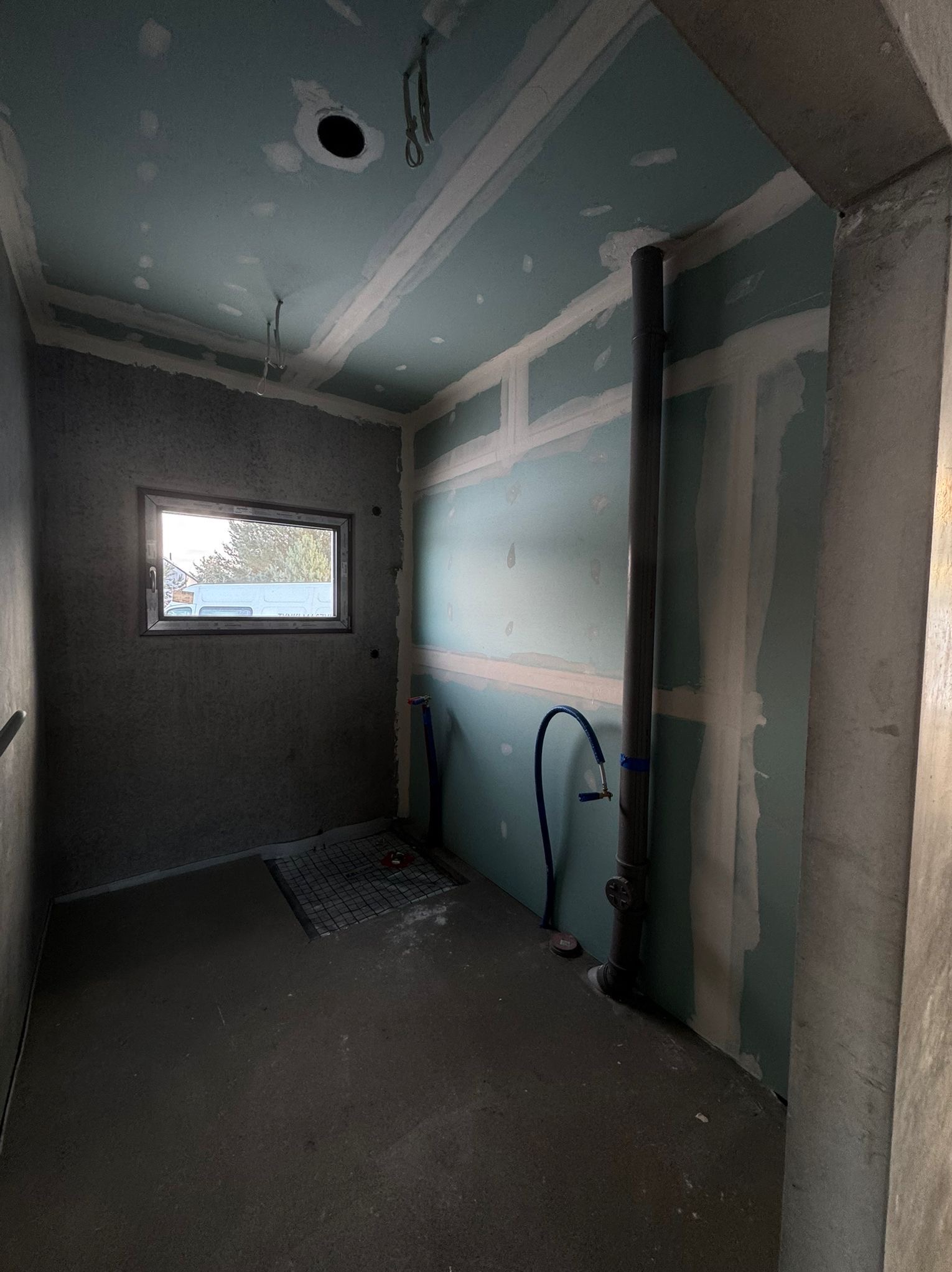 Surowe wnętrze w trakcie wykańczania: ściany z płyt karton-gips, widoczne instalacje, okno z widokiem na zewnątrz. Etap prac przed ostatecznym wykończeniem.