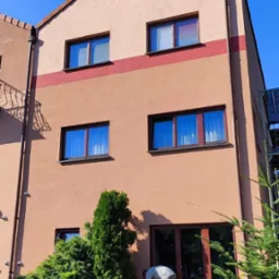 Dwupiętrowy budynek z beżową elewacją i brązowymi oknami pod błękitnym niebem; w tle widoczna zieleń, a na pierwszym planie osoba sprzątająca przed wejściem.