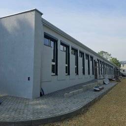 F.H.U. Partner - Długi budynek po tynkowaniu z szarymi ścianami, czarnymi ramami okiennymi i brukowanym chodnikiem. Widoczny fragment nieutwardzonego terenu.