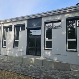 F.H.U. Partner - Jasnoszara elewacja budynku z ciemnymi ramami okiennymi i drzwiami. Widoczny podjazd dla niepełnosprawnych z kostki brukowej. Nowoczesny design budynku.