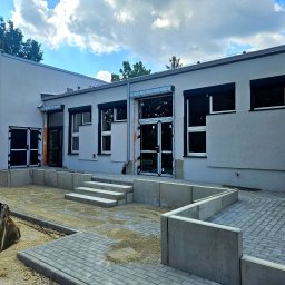 F.H.U. Partner - Elewacja budynku po tynkowaniu, widoczne okna z roletami, schody wejściowe, kostka brukowa i łyżka koparki. Szara kolorystyka, niebo z chmurami w tle.