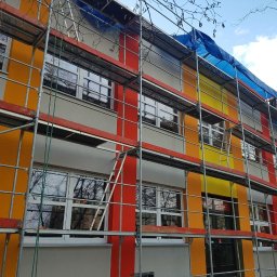 F.H.U. Partner - Budynek w rusztowaniach z pomalowaną elewacją w pasy: żółte, pomarańczowe i czerwone. Na górze plandeka i pracownik w kamizelce.