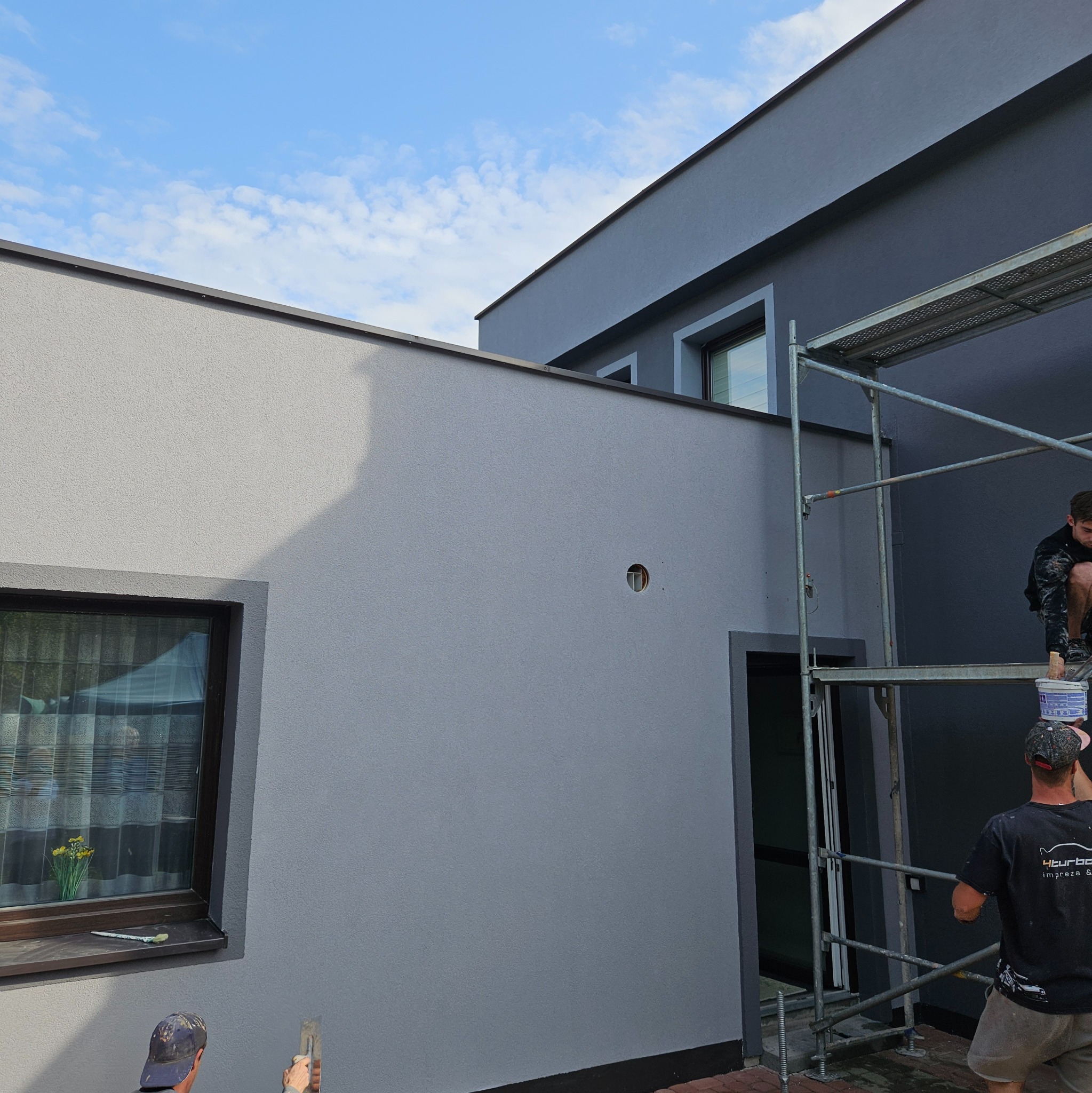 Dwóch pracowników na rusztowaniu podczas prac elewacyjnych nowoczesnego domu w odcieniach szarości. Jeden z nich trzyma wiadro z farbą, drugi przygotowuje się do malowania.