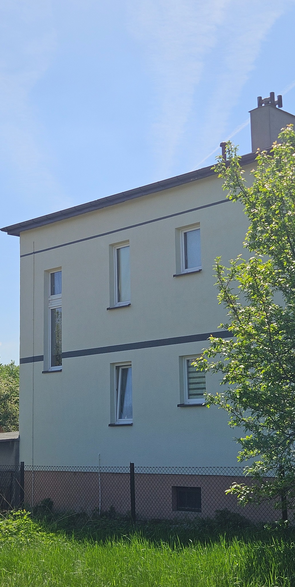 Jasna elewacja dwukondygnacyjnego budynku z ciemnymi pasami, widok z poziomu trawy, z fragmentem drzewa po prawej stronie. Widoczne okna i komin.