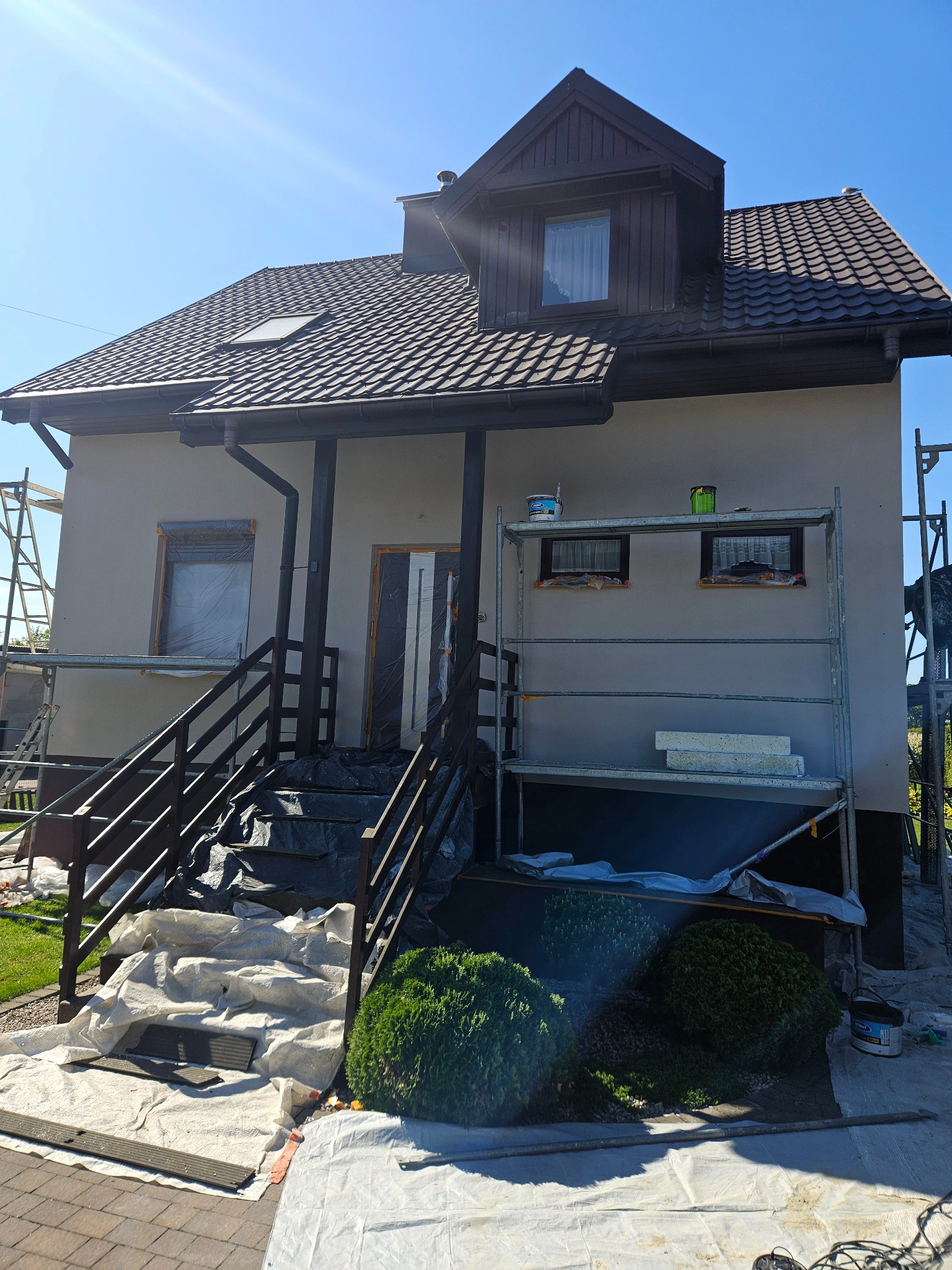 Dom jednorodzinny w trakcie prac elewacyjnych: rusztowania, zabezpieczone okna, widoczne materiały budowlane. Ujęcie z dołu, w słoneczny dzień.