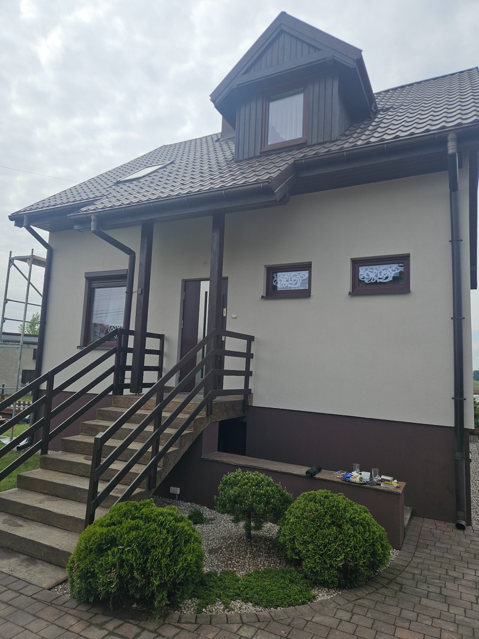 Wykończona elewacja domu jednorodzinnego z brązowymi akcentami: dach, okna, rynny. Świeżo otynkowana, jasna fasada z widocznymi schodami wejściowymi.