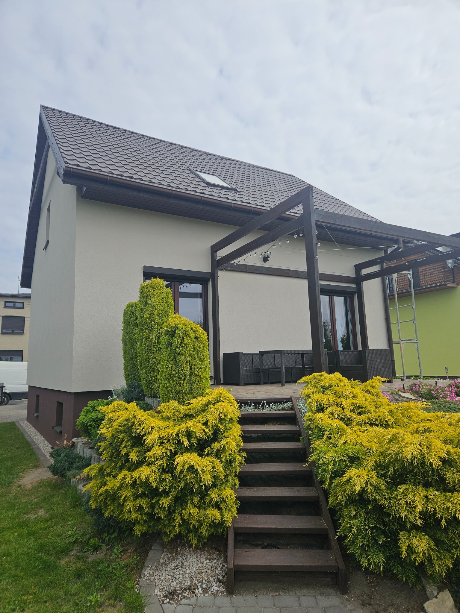 Elegancki dom po renowacji w Rogoźniku, z jasną elewacją i brązowymi detalami, widok z dołu, z zadbanym ogrodem i schodami na taras.