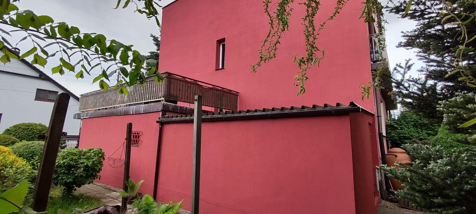 Różowa elewacja budynku z balkonem, widok z dołu. Fragmenty zieleni w kadrze, małe okno na ścianie, ciemna dachówka. Ujęcie z bliska, bez detali.