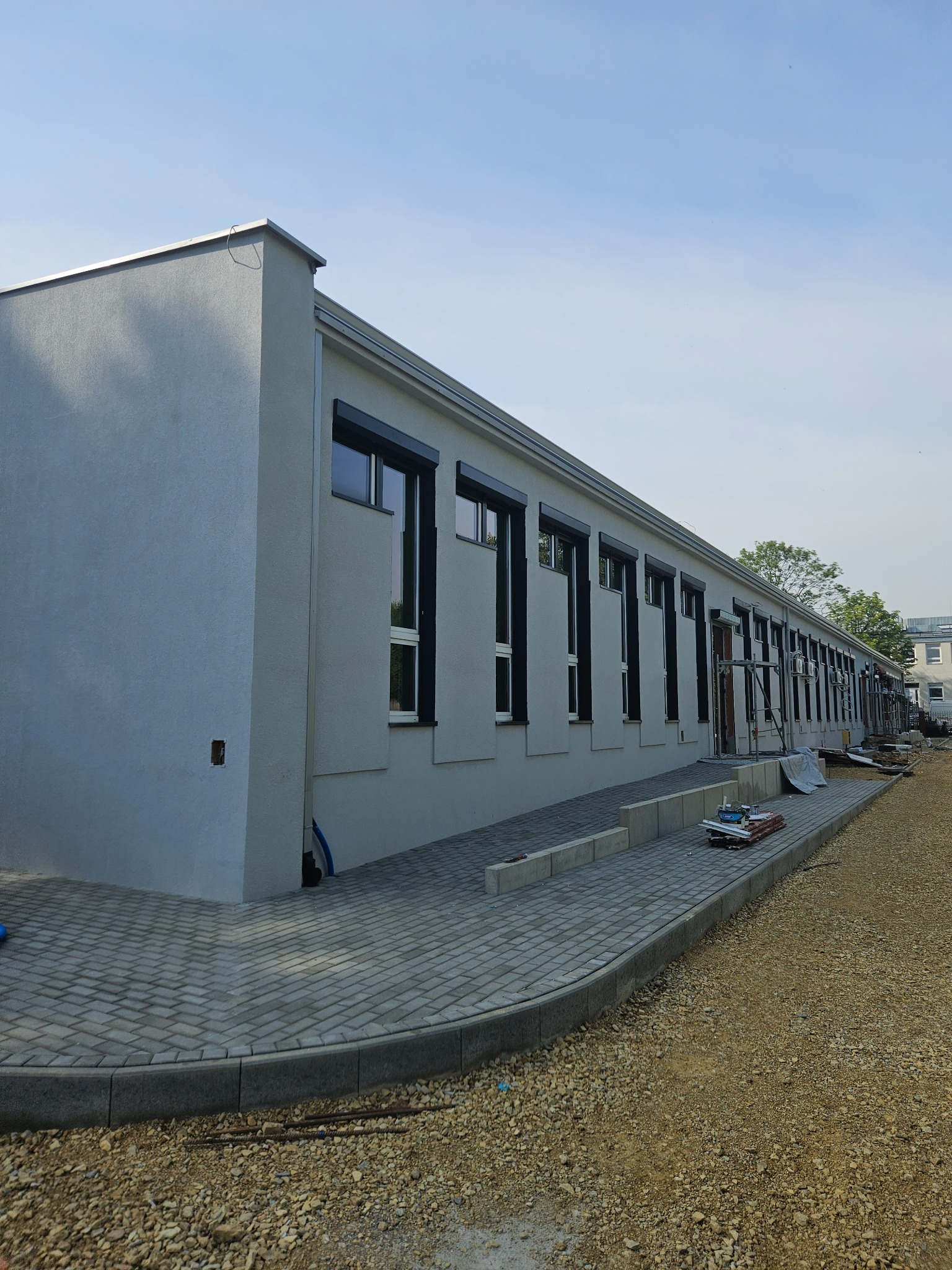 Długi budynek po tynkowaniu z szarymi ścianami, czarnymi ramami okiennymi i brukowanym chodnikiem. Widoczny fragment nieutwardzonego terenu.