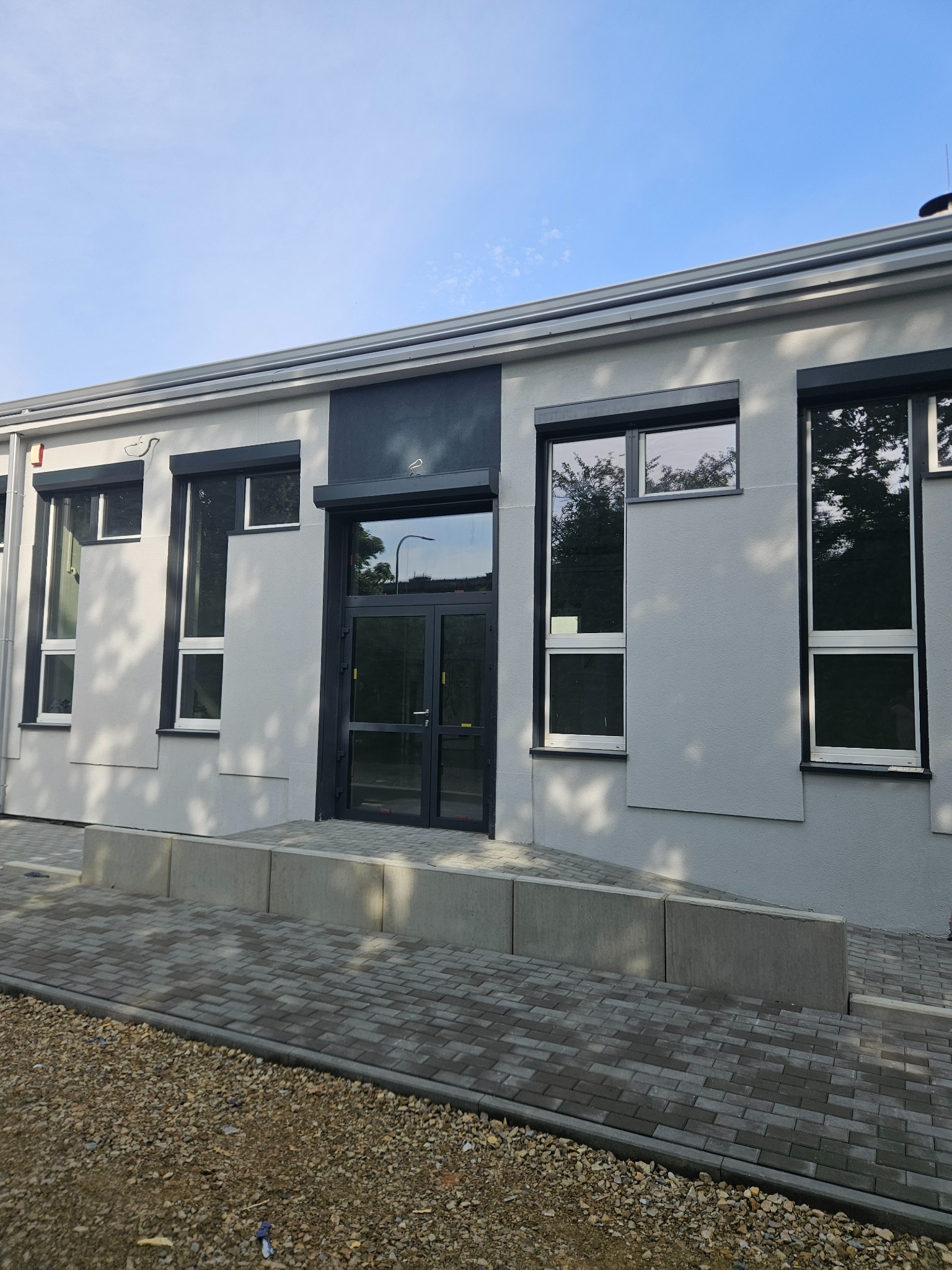 Jasnoszara elewacja budynku z ciemnymi ramami okiennymi i drzwiami. Widoczny podjazd dla niepełnosprawnych z kostki brukowej. Nowoczesny design budynku.