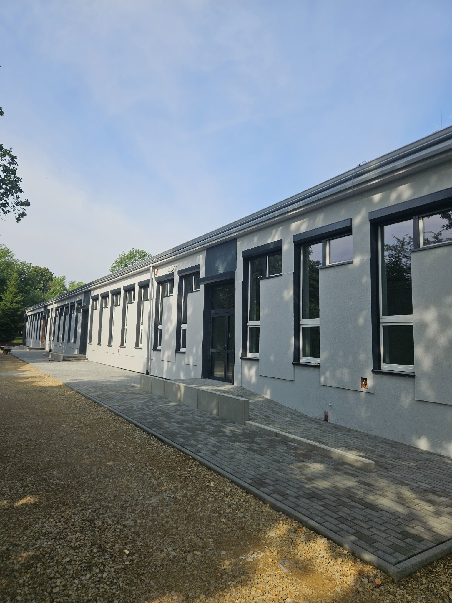 Odnowiona elewacja budynku z szarym tynkiem, czarnymi ramami okiennymi i kostką brukową. Widok z zewnątrz, zieleń drzew w tle.