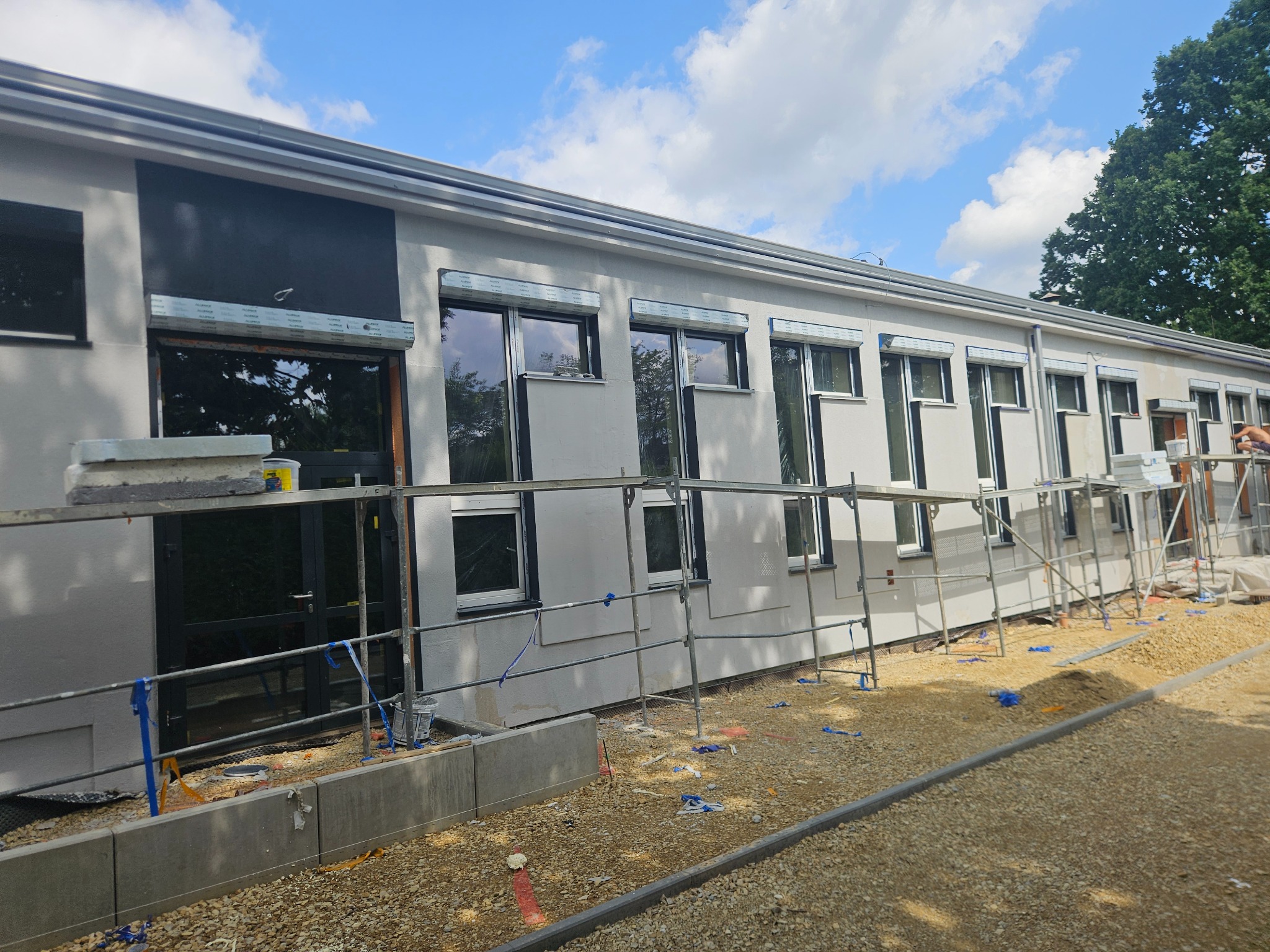 Elewacja budynku po tynkowaniu w Rogoźniku. Widoczne rusztowania i okna z roletami. Fragment pomalowanej ściany na czarno. Prace w toku, teren wokół wysypany żwirem.