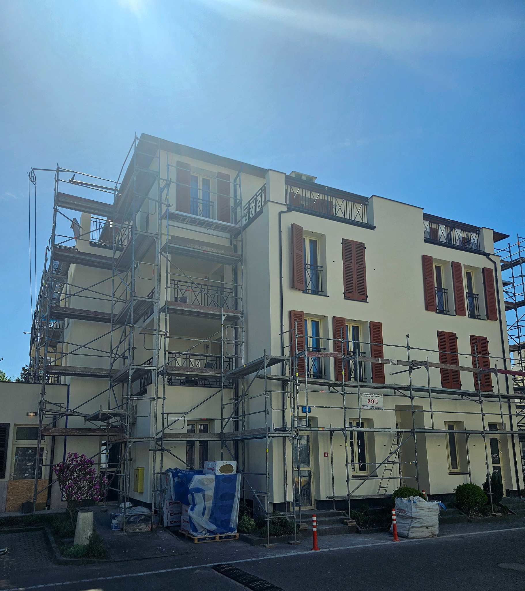 Biała elewacja budynku z brązowymi okiennicami w trakcie prac; widoczne rusztowania i materiały budowlane. Słoneczny dzień.