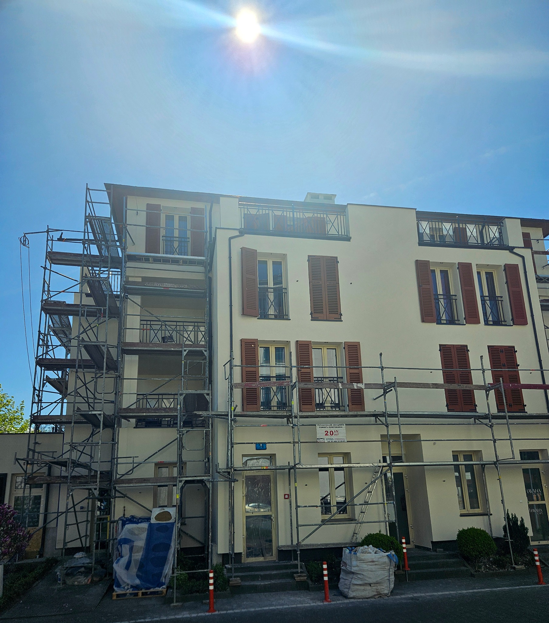 Elewacja budynku w trakcie prac. Widoczne rusztowania, biała fasada z brązowymi okiennicami. Słoneczny dzień, fragment nieba.