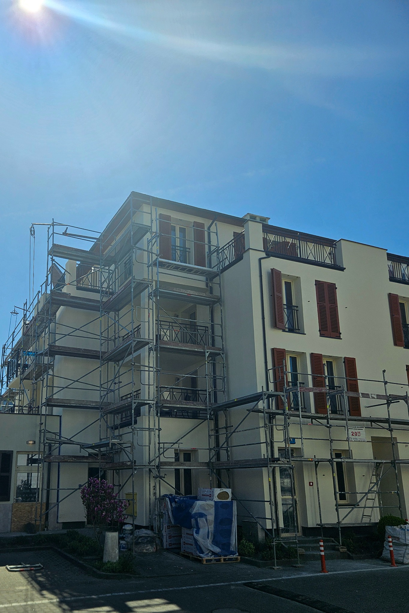 Biały budynek mieszkalny z brązowymi okiennicami w trakcie prac elewacyjnych, z rusztowaniem i materiałami budowlanymi na pierwszym planie.