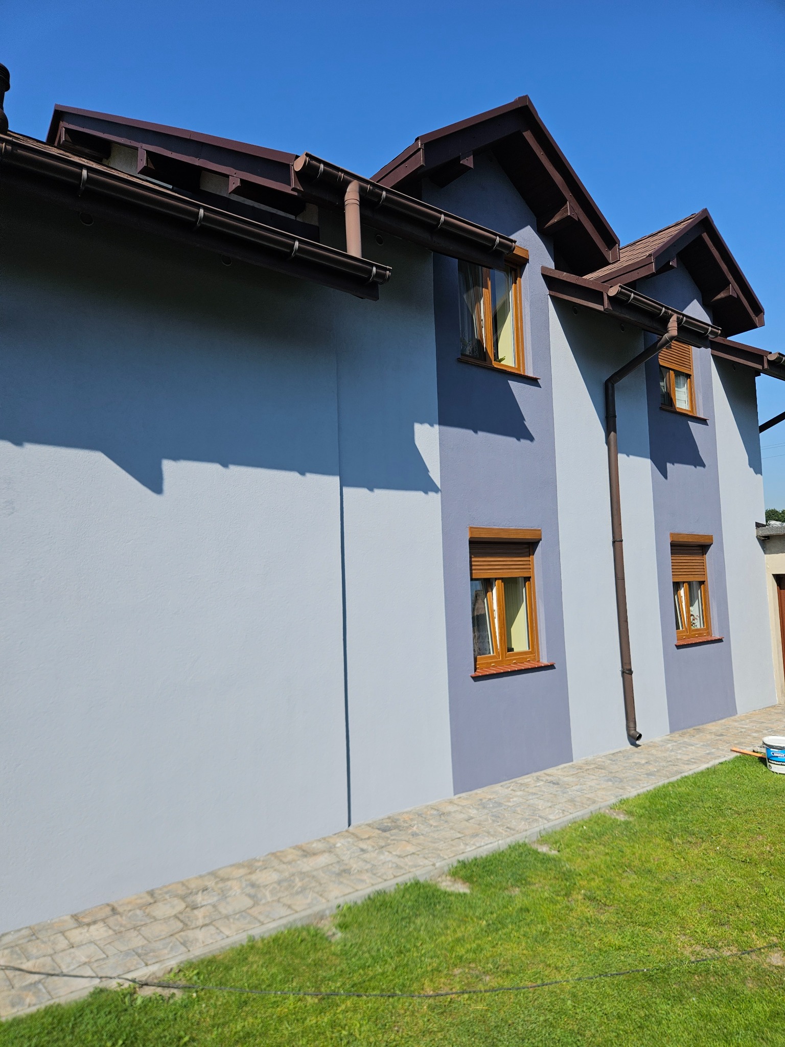 Dwukolorowa elewacja domu z brązowymi rynnami i oknami z roletami, widoczna z poziomu trawnika. Świeżo położony tynk. Na trawie stoi wiadro z farbą.