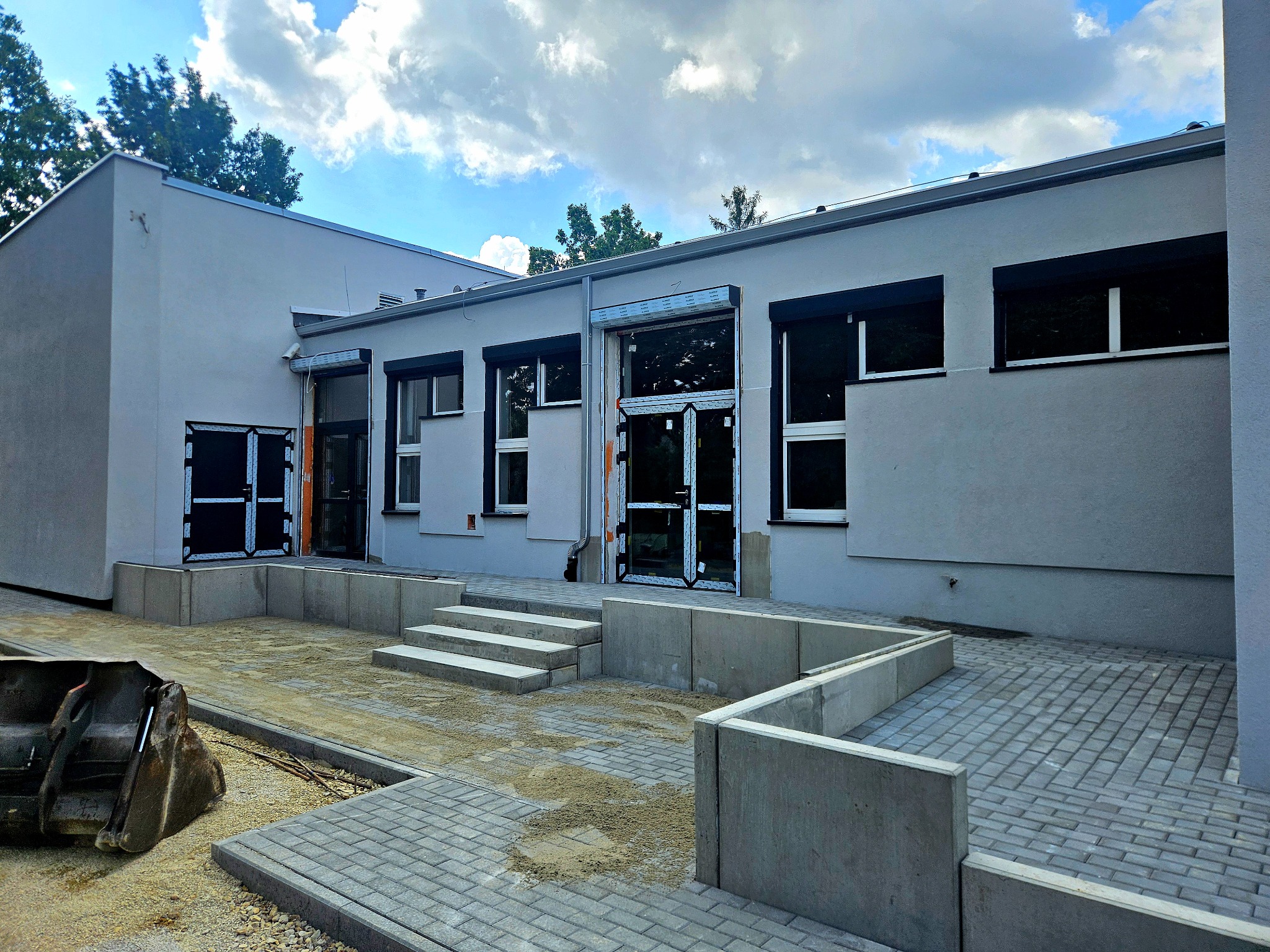 Jasnoszara elewacja budynku po remoncie z czarnymi roletami i ramami okiennymi. Kostka brukowa przed wejściem i łyżka koparki w rogu kadru.