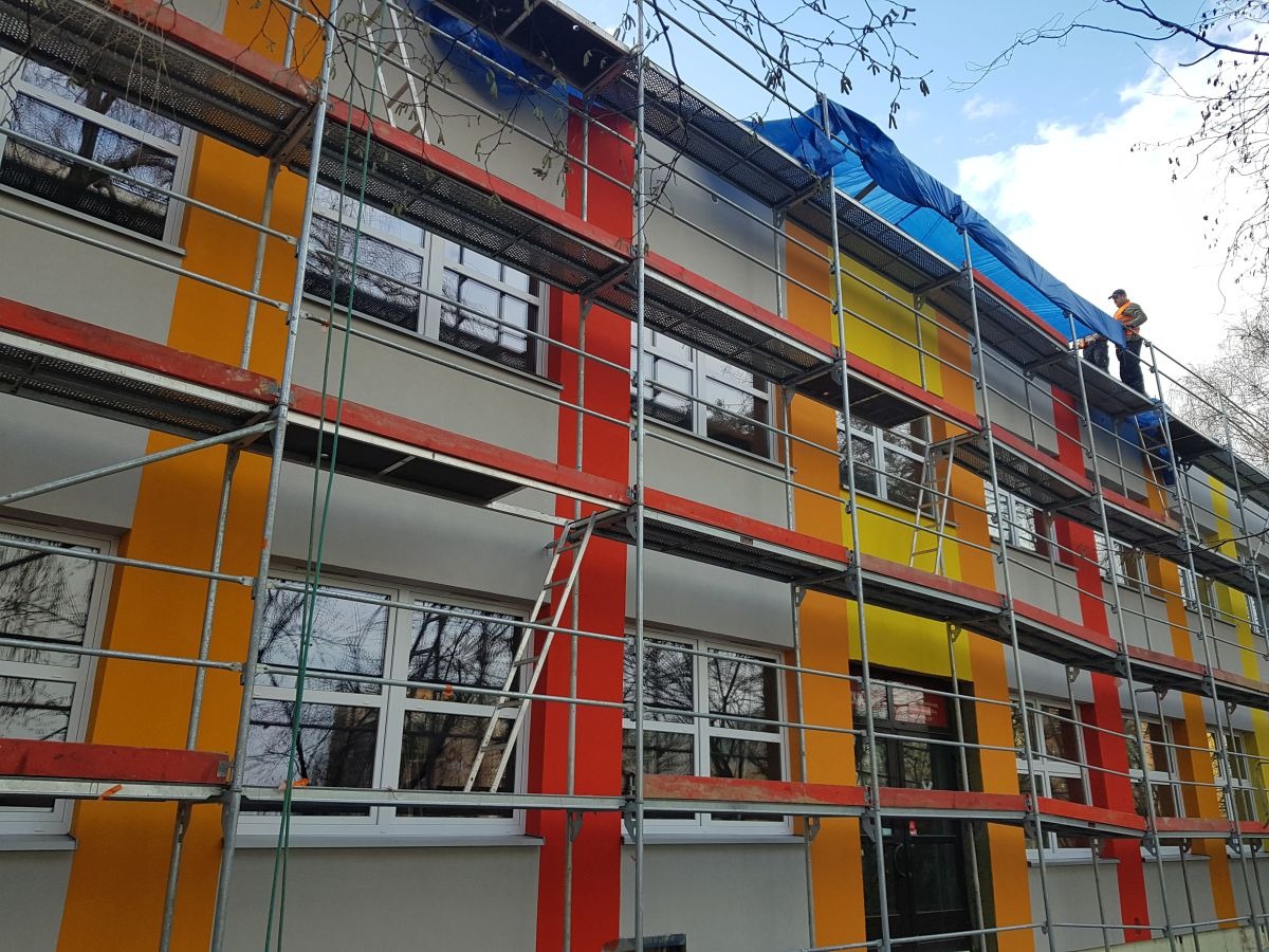 Budynek w rusztowaniach z pomalowaną elewacją w pasy: żółte, pomarańczowe i czerwone. Na górze plandeka i pracownik w kamizelce.