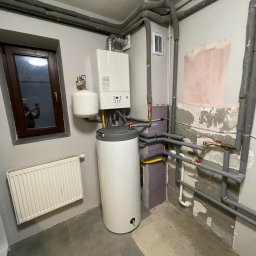 Serwis pieców gazowych KANIGOWSKI Montaż, Naprawa i Przegląd pieca gazowego - Kompaktowa kotłownia w piwnicy: wiszący piec gazowy z zasobnikiem c.w.u. oraz orurowanie z izolacją termiczną. W oknie odbicie osoby robiącej zdjęcie.