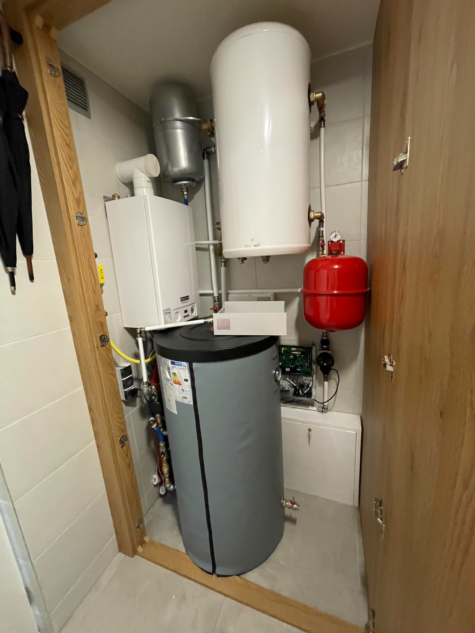 Kompaktowa kotłownia w szafie: wiszący piec gazowy, biały zasobnik CWU, szary zbiornik buforowy i czerwone naczynie przeponowe, całość otoczona białą glazurą i drewnianą konstrukcją.