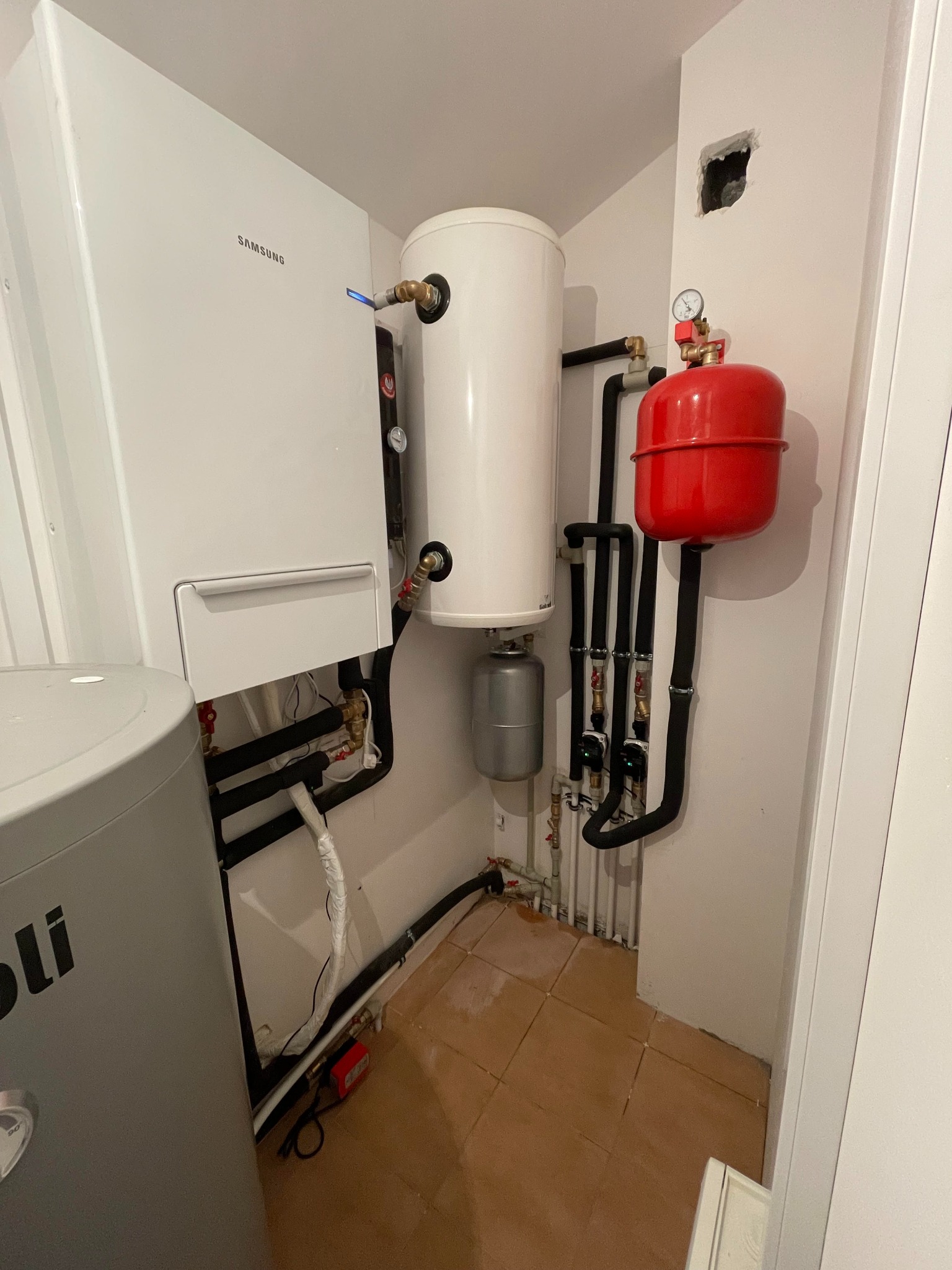 Nowoczesny system grzewczy z piecem gazowym Samsung, zbiornikiem akumulacyjnym i czerwonym naczyniem wzbiorczym w pomieszczeniu technicznym w Michałowie-Grabinie. Widoczne rury i przyłącza instalacji.
