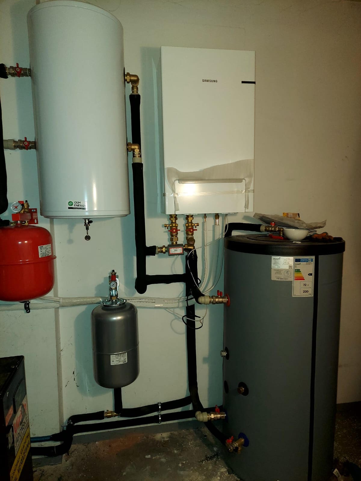 Instalacja centralnego ogrzewania z piecem gazowym Samsung, zbiornikiem CWU i naczyniami przeponowymi, widoczne rury w izolacji i zawory odcinające.