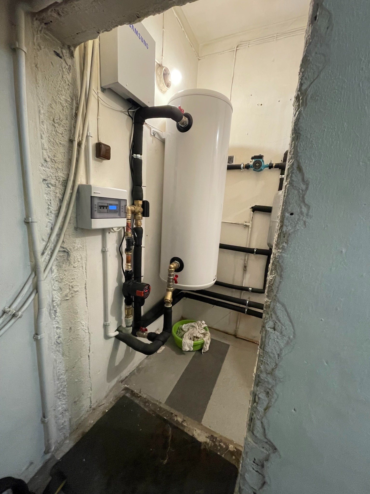 Biały, pionowy zbiornik ciepłej wody podłączony do instalacji grzewczej w piwnicy, widoczne rury, zawory, regulator temperatury i osprzęt Samsung.