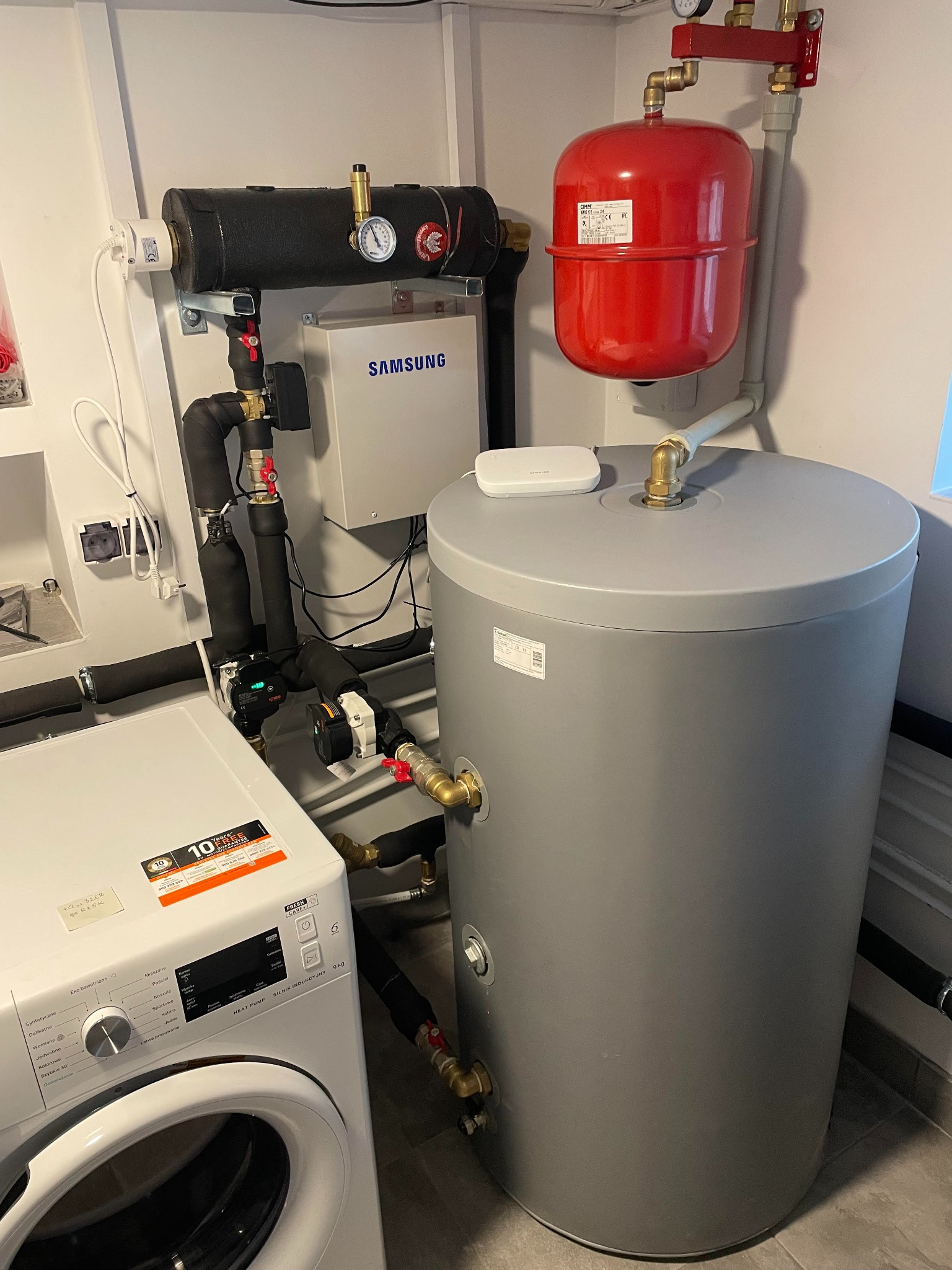 Wnętrze kotłowni z szarym bojlerem, czerwonym zbiornikiem wyrównawczym CIMM, czarnymi rurami izolowanymi, białą pralką z naklejką 10 lat gwarancji i skrzynką sterującą Samsung na ścianie.
