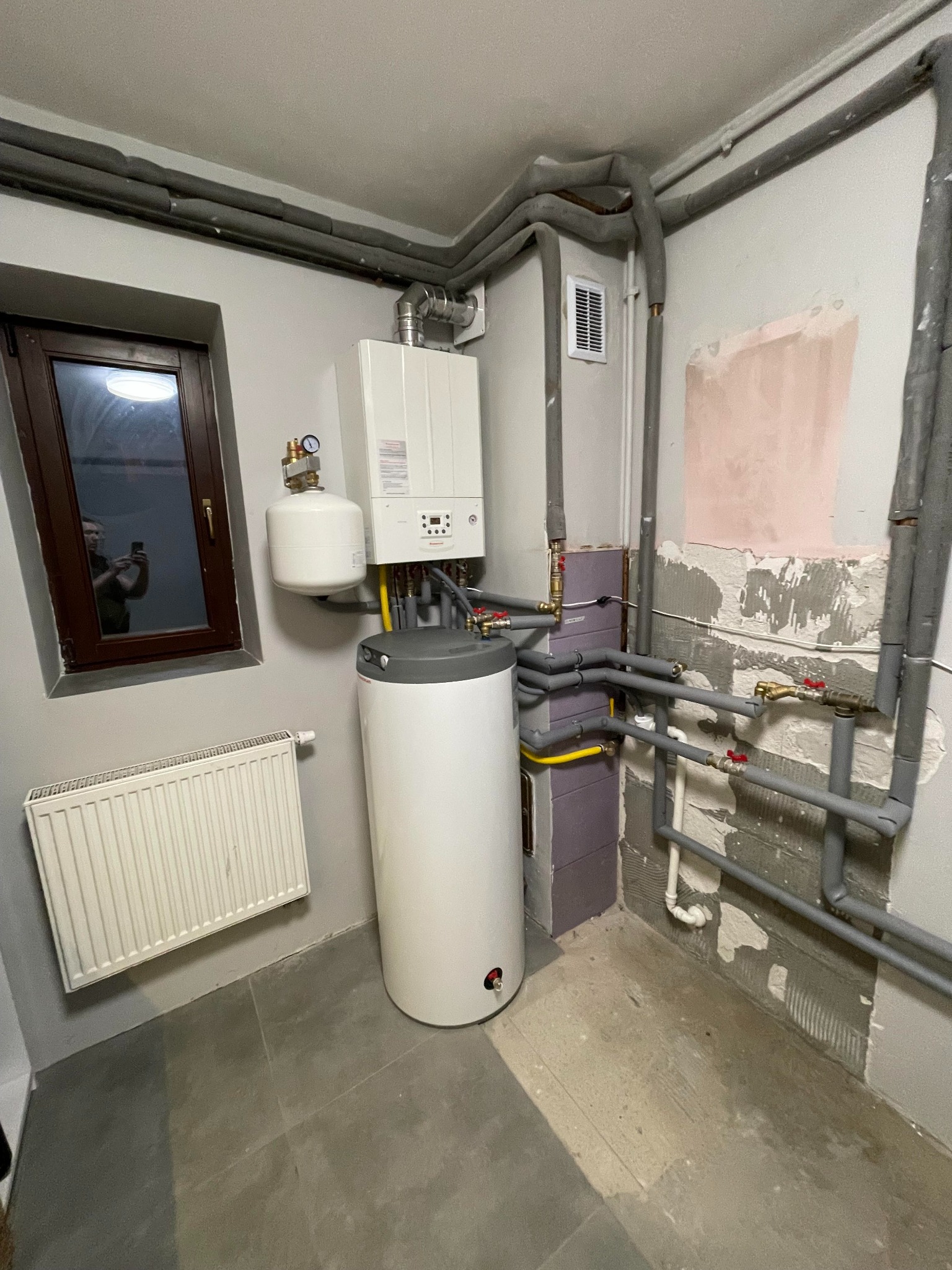 Kompaktowa kotłownia w piwnicy: wiszący piec gazowy z zasobnikiem c.w.u. oraz orurowanie z izolacją termiczną. W oknie odbicie osoby robiącej zdjęcie.