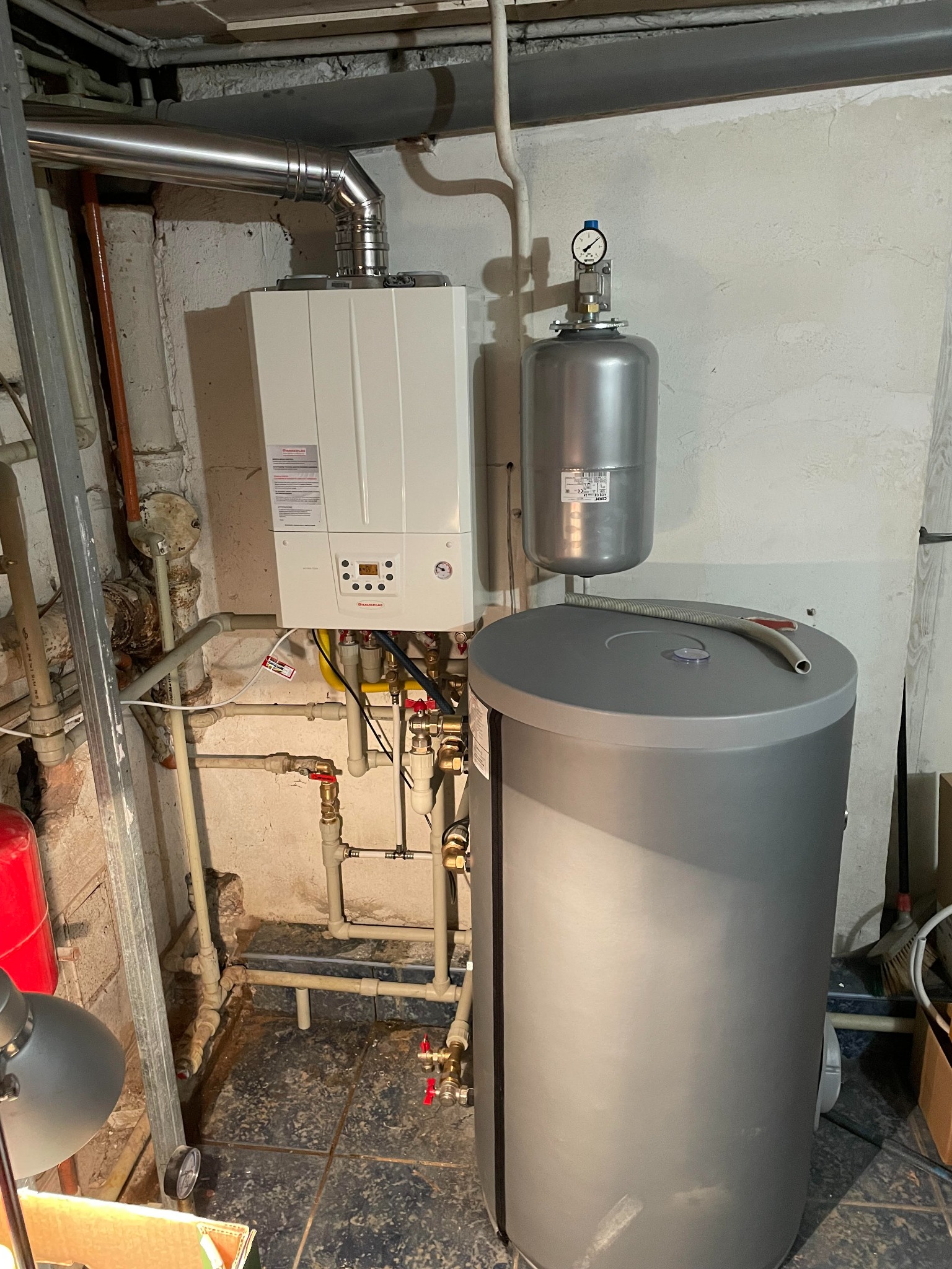 Biały piec gazowy kondensacyjny zamontowany na ścianie w piwnicy, z podłączonymi rurami, zbiornikiem wyrównawczym i zasobnikiem ciepłej wody; widoczne liczne przyłącza i zawory.