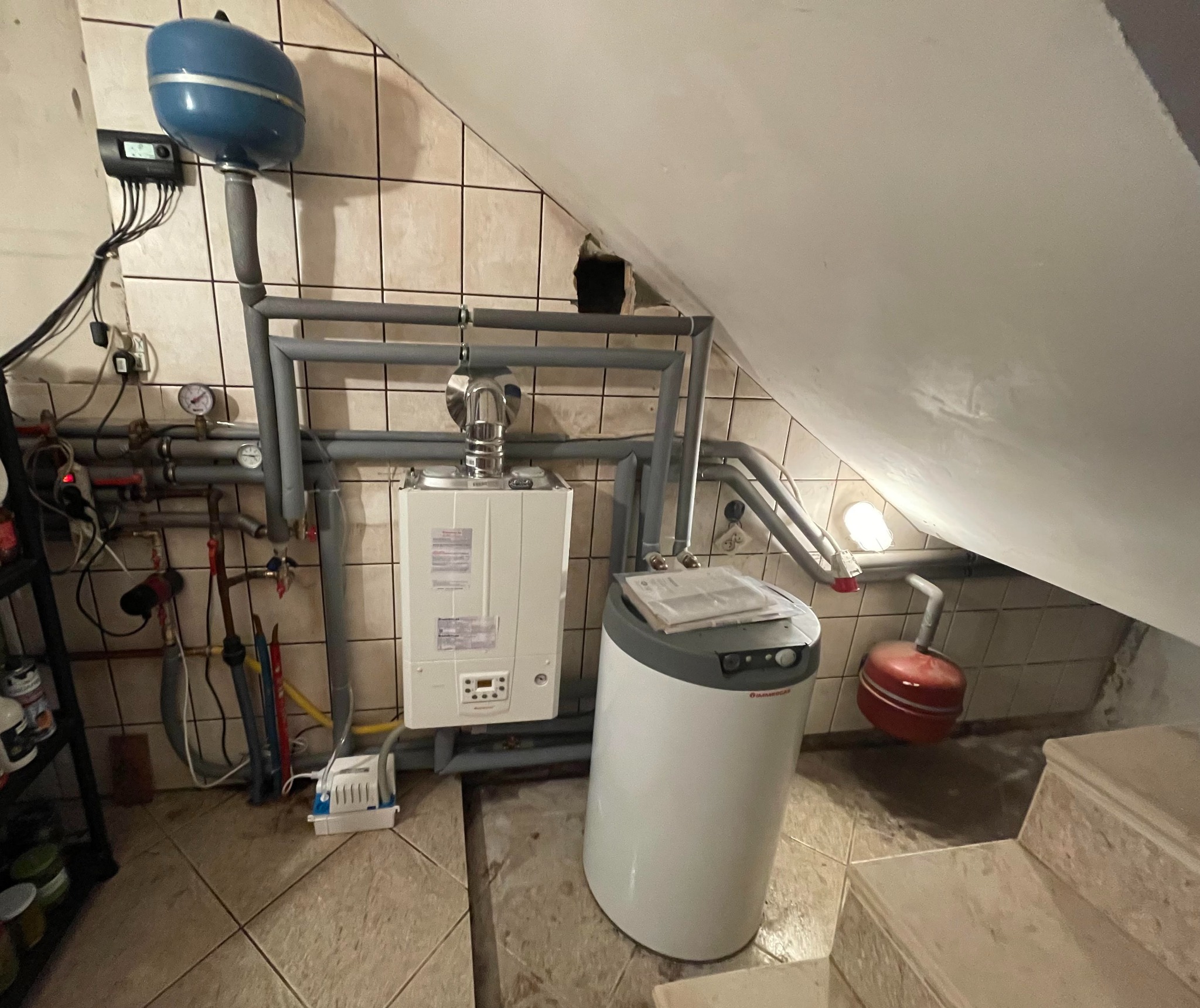 Ukończona instalacja pieca gazowego w pomieszczeniu z beżowymi płytkami, widoczne rury, zbiornik wyrównawczy, sterownik, oraz piec Immergas z podłączonym zasobnikiem wody. W tle stary otwór...