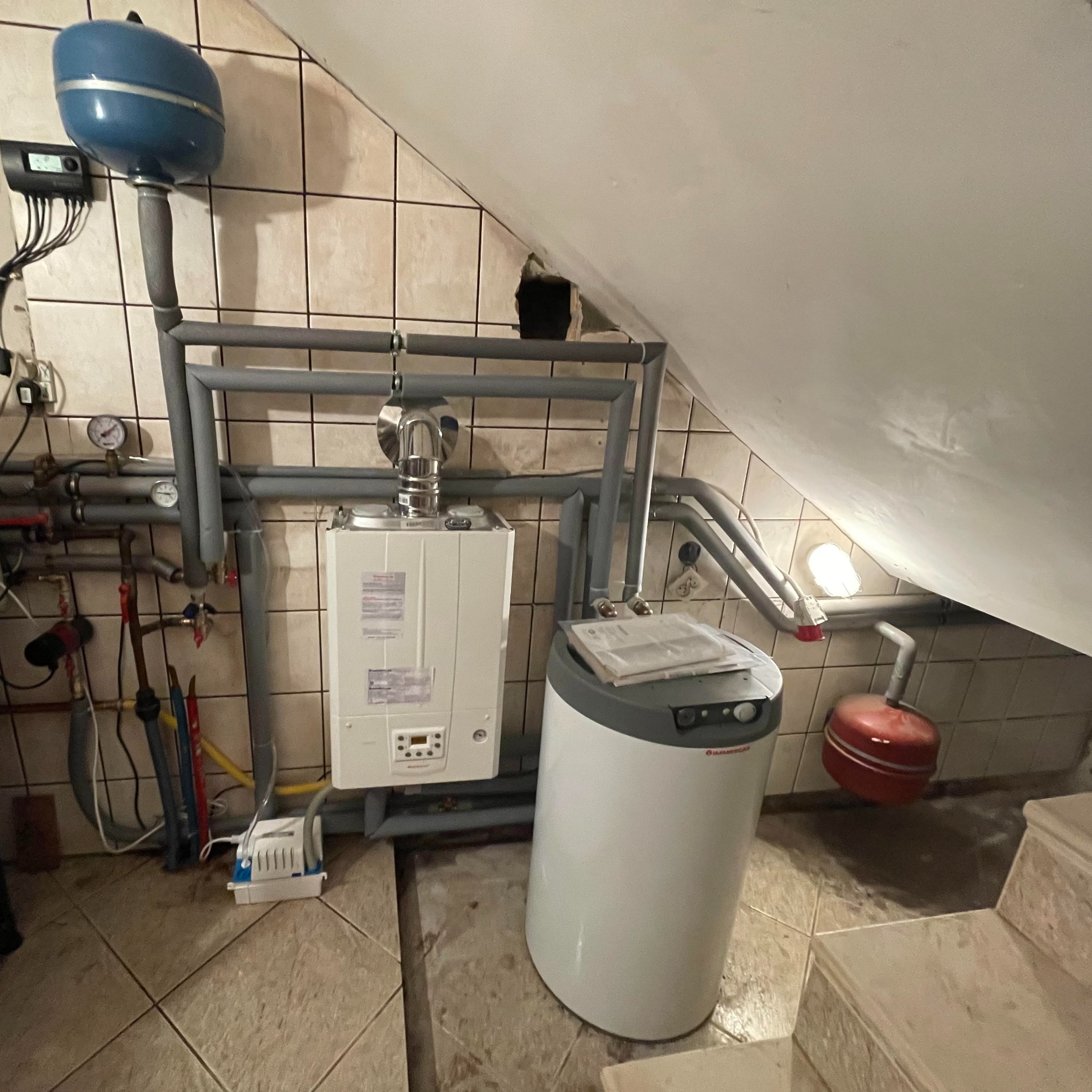 Kocioł gazowy wiszący na ścianie z ceramicznych płytek, obok biały zasobnik ciepłej wody, instalacja z szarych rur, mała kotłownia na poddaszu.