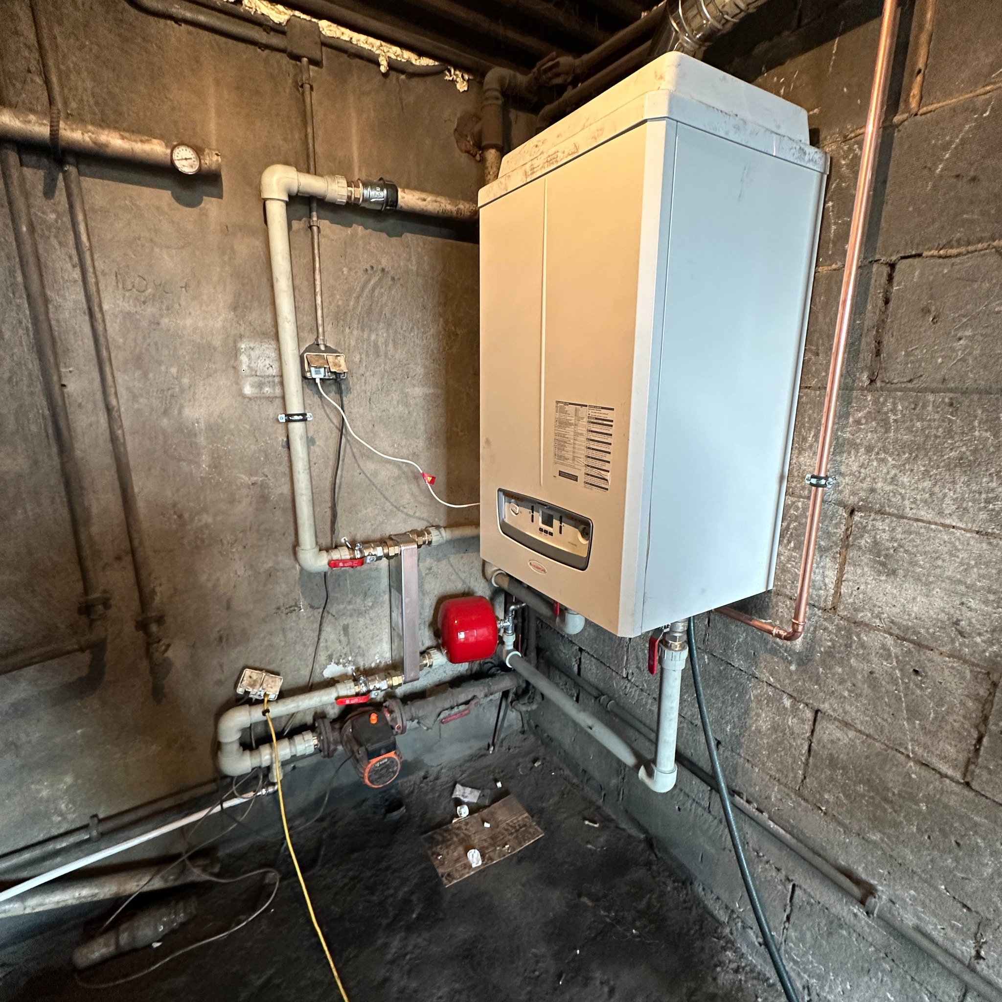 Biały piec gazowy zamontowany na szarej, surowej ścianie z widocznymi rurami miedzianymi i plastikowymi oraz czerwonym zbiornikiem wyrównawczym.