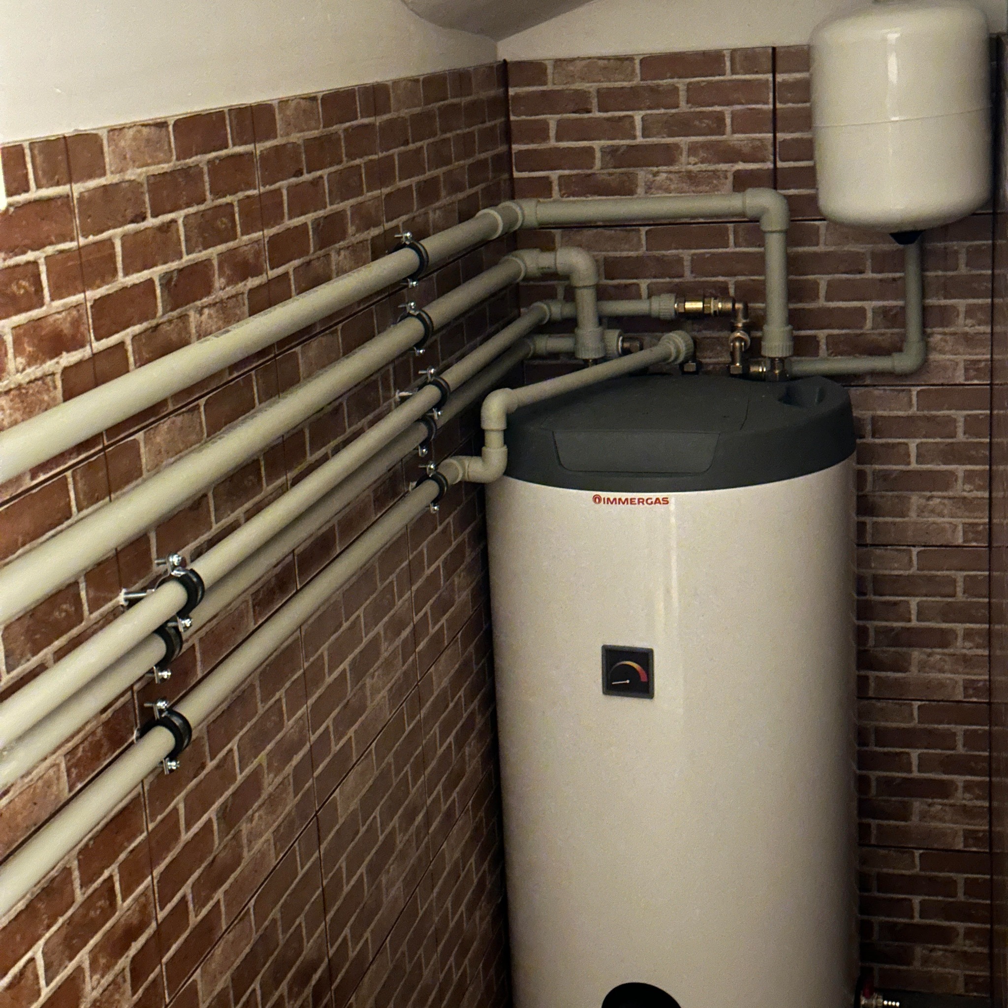 Biały piec gazowy Immergas z szarą górą, widoczne rury, zamocowany do ściany wyłożonej cegłą, zbiornik wyrównawczy u góry.