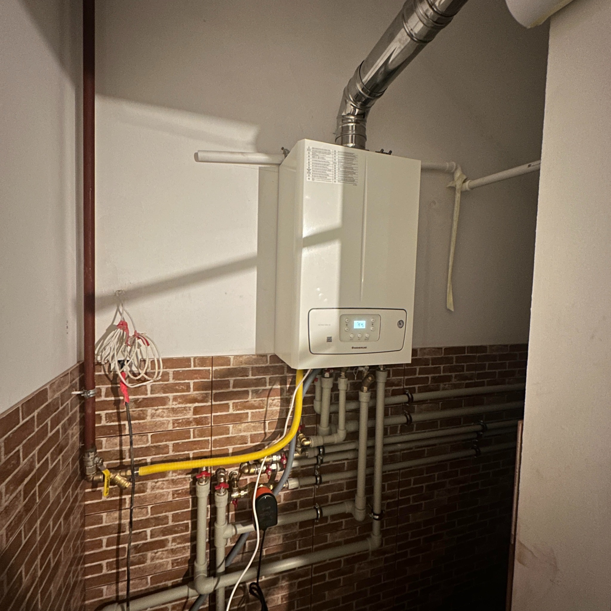 Nowoczesny piec gazowy wiszący na ścianie z cegły, z widocznymi rurami doprowadzającymi gaz i wodę, oraz przewodem odprowadzającym spaliny.